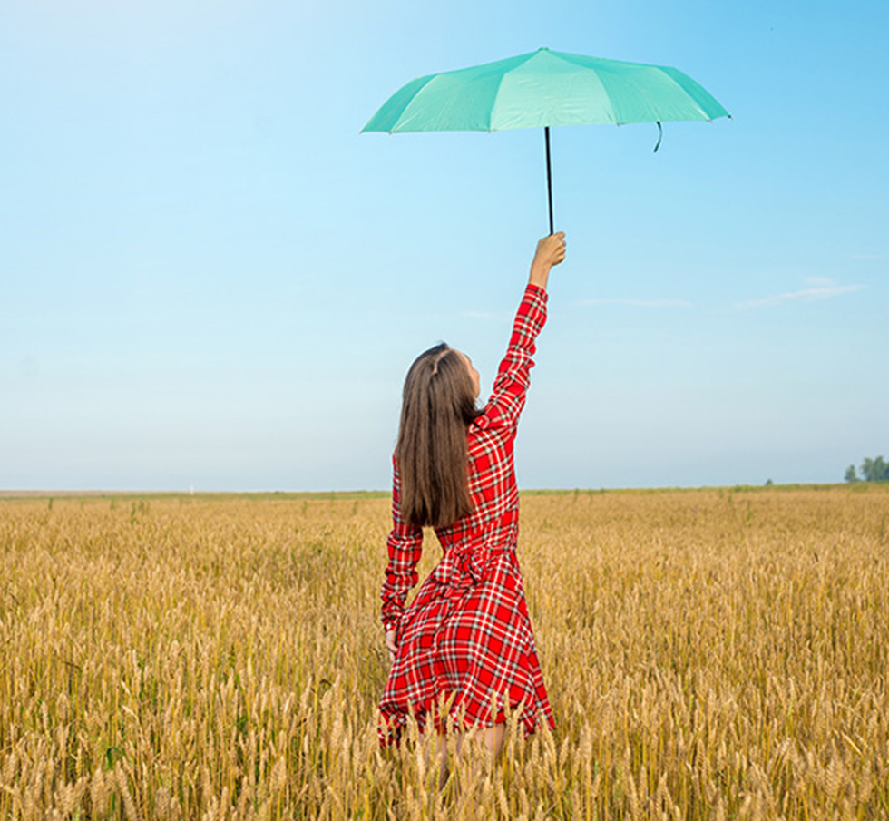 A woman in a red plaid dress holding a green umbrella while standing in a wheat field.